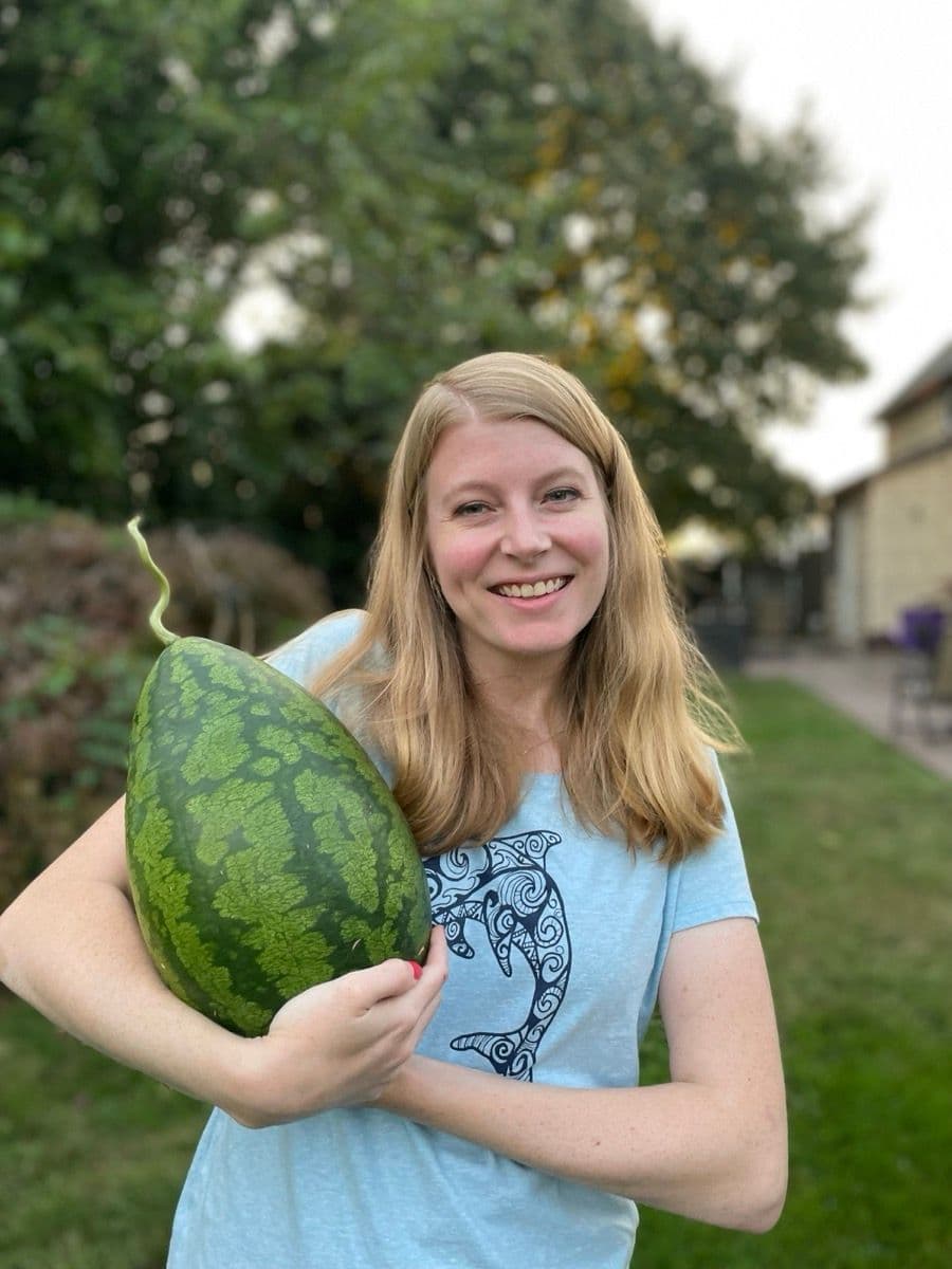 Liz smiling and holding a freshly harvested watermelon in her backyard garden